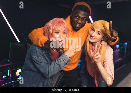 Multirassische junge, fröhliche Freunde, die nahe beieinander stehen, posieren in einem dunklen Spielzimmer vor der Kamera, mit Neonlichtern, die Frieden zeigen und mit Daumen nach oben Zeichen. Hochwertige Fotos Stockfoto