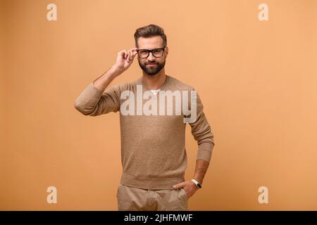 Charismatischer bärtiger kaukasischer gutaussehender Mann, isoliert auf beigem Hintergrund stehend und schaut auf die Kamera, männlicher Unternehmer in legerer Kleidung, der stilvolle Brillen anpasst Stockfoto