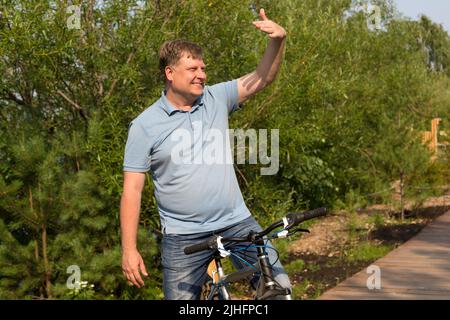 Ein lächelnder Erwachsener auf einem Fahrrad ruft einen Freund mit einer Handbewegung an. Stockfoto