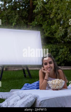 Vertikale Ansicht einer jungen Frau, die lächelnd auf die Kamera blickt, mit einer Projektionsleinwand im Hintergrund und einigen Popcornen Stockfoto