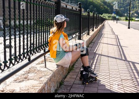 Eine junge Frau in einer gelben Sweatshirt-Mütze in Schutzausrüstung und Rollschuhe sitzt auf dem Damm und schaut nach dem Rutschengehen entspannt in die Ferne Stockfoto