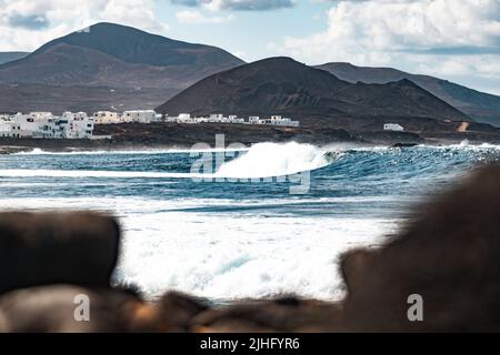 Wilde felsige Küste des Surfspots La Santa Lanzarote, Kanarische Inseln, Spanien. Surfer reiten eine große Welle in felsiger Bucht, Vulkan Berg im Hintergrund. Stockfoto