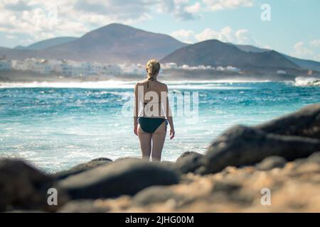 Weibliche Touristen am wilden felsigen Strand und der Küste des Surfspots La Santa Lanzarote, Kanarische Inseln, Spanien. La Santa Dorf und Vulkan Berg in Stockfoto