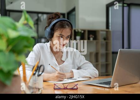 Glückliche junge asiatische Mädchen mit kabellosen Kopfhörern Blick auf Laptop-Bildschirm, Lesen hören Online-Kurse, Lernen aus der Ferne von zu Hause wegen Stockfoto