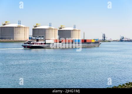 An klaren Sommertagen segelt ein Containerschiff an LNG-Tanks in einem Hafen vorbei Stockfoto