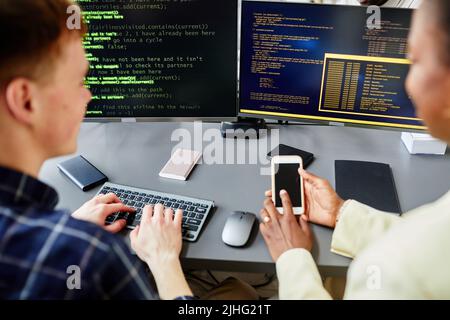 Rückansicht eines afrikanischen Mädchens, das auf das Smartphone zeigt und sich mit ihrer Kollegin am Tisch mit Computern berät Stockfoto