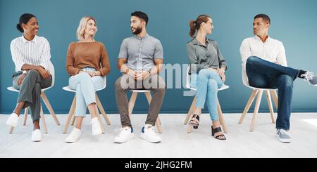 Eine vielfältige Gruppe von selbstbewussten Geschäftsleuten, die auf Stühlen im Warteraum vor blauem Hintergrund sitzen. Glückliche hoffnungsvolle Kandidaten und Bewerber Stockfoto