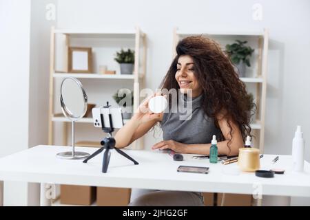 Happy cute gebräunt schöner lockiger lateinischer Beauty-Blogger in Casual Top erzählt über Gesichtscreme im Heim-Interieur. Space Mockup Banner kopieren. Einflussnehmer Stockfoto