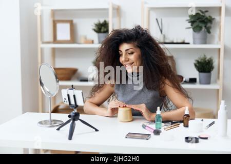 Fröhlich gebräunt schöner lockiger lateinischer Beauty-Blogger in Casual Top erzählt von Körpercreme im Heim-Interieur. Space Mockup Banner kopieren. Influencer-Rekord Stockfoto