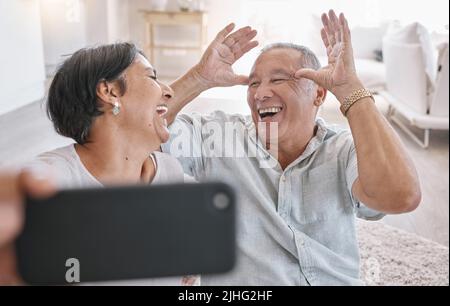 Nahaufnahme eines liebevollen Seniorenpaares, das Selfies macht, während es sich im Wohnzimmer zu Hause entspannt. Gemischtes Rennpaar, das Spaß hat, Fotos in der zu machen Stockfoto