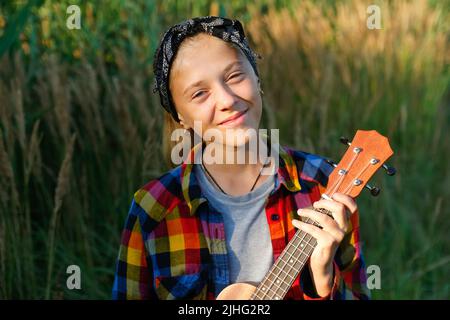 Defokussieren Mädchen mit Gitarre. Teen Mädchen zu Fuß auf Natur Hintergrund. Kleines Mädchen draußen. Grüne Wiese. Generation z. Herbst. Bandana. Lifestyle Stockfoto