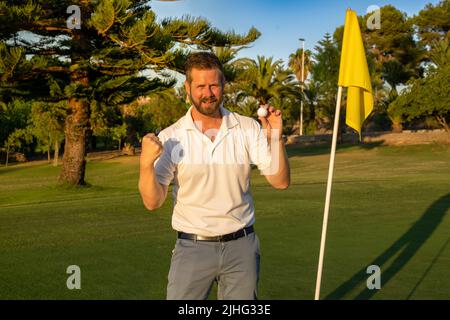 Glücklicher Golfspieler, der den Ball hält, macht eine erfolgreiche Geste am Golfclub. Stockfoto