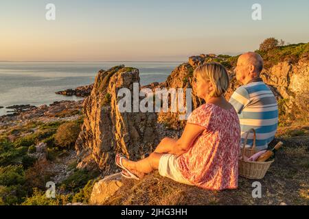 Das Ehepaar Hovs Hallar, Skåne, Schweden, sitzt auf einer Klippe und genießt den Sonnenuntergang bei einem Picknick mit Wein und Brot Stockfoto