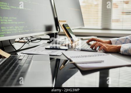 Nahaufnahme einer weiblichen Programmiererin, die vom Smartphone Code auf die Tastatur eingibt, während sie am Tisch vor Computermonitoren im Büro sitzt Stockfoto