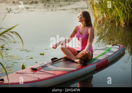 Schönes Mädchen, das in Lotusposition mit geschlossenen Augen sitzt und auf dem Wassersportbrett meditiert. Eine Frau auf einem Sup und eine Atmosphäre der Entspannung auf Th Stockfoto