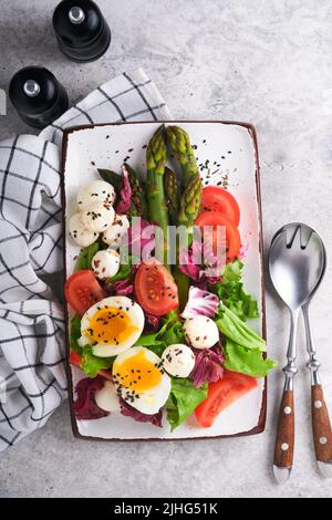 Spargel, Tomaten, Salat, Mozzarella, schwarzer Sesam, Flachs, Ölolivsalat und weich gekochtes Ei auf rechteckiger Keramikplatte auf grauem Betontisch Bac Stockfoto