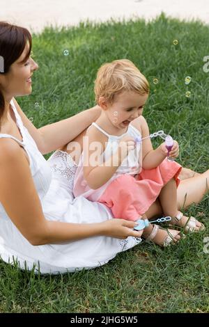 Glückliche Mutter, die ihre Tochter anschaut, die auf dem Rasen mit Seifenblasen spielt Stockfoto