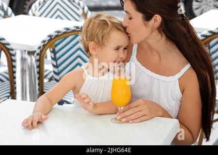 Mutter küsst lächelnde Kleinkindtochter in der Nähe von Orangensaft im Café im Freien in Valencia Stockfoto