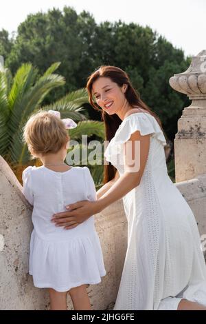 Glückliche Frau im Sommerkleid umarmte Tochter auf dem Balkon des Steingebäudes in Valencia Stockfoto