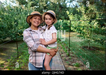 Fröhliche Frau, Mutter hält ihre kleine Tochter auf den Armen und steht in Reihen von Setzlingen auf dem Bio-Bauernhof zusammen Stockfoto
