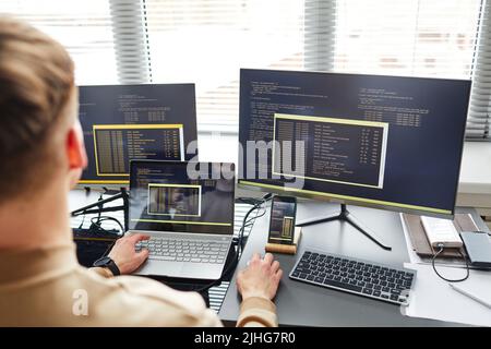Rückansicht eines jungen IT-Spezialisten, der Laptop mit Computer verbindet, während er Daten auf der Tastatur am Arbeitsplatz eingibt Stockfoto