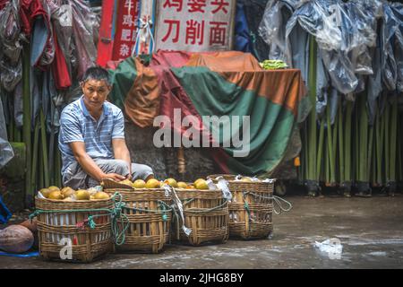 Zigong, China - Juli 2019 : Chinesischer Mann, der auf dem Bürgersteig sitzt und Gemüse verkauft, das in Korbkörben aus Bambus gelagert ist, Stadt Zigong, Provinz Sichuan Stockfoto
