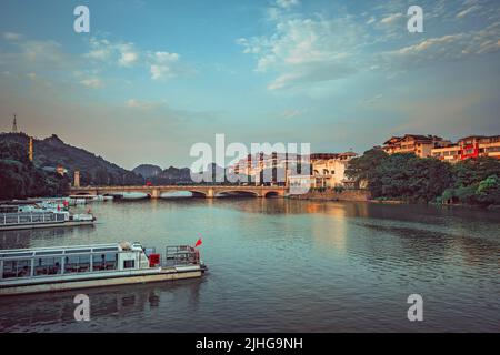 Guilin, China - August 2019 : Lastkähne dockten am Flussufer an, mit einer gewölbten Straßenbrücke über den prächtigen Li Fluss im Hintergrund, Stadt Guilin, Guangxi Stockfoto