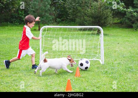 Kid Boy in rot-weißem Trikot spielt Fußball (Fußball) mit seinem Hund auf dem Trainingsgelände Stockfoto