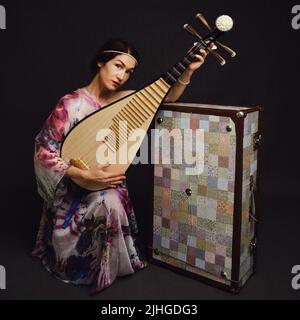 Eine glückliche Musikerin in asiatischen Kleidern auf einem schwarzen Hintergrund im Studio. Eine lächelnde Frau mit einem Saiteninstrument aus Asien auf einem dunklen Backgrou Stockfoto