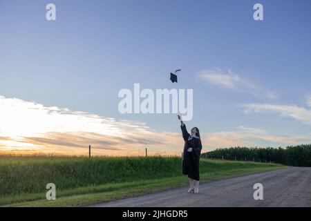 Junge Frau in Abschlusskleid, die ihre Mütze und ihr Mörtelbrett in die Luft wirft, mit einem blauen Himmel im Hintergrund Stockfoto