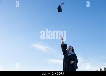 Junge Frau in Abschlusskleid, die ihre Mütze und ihr Mörtelbrett in die Luft wirft, mit einem blauen Himmel im Hintergrund Stockfoto