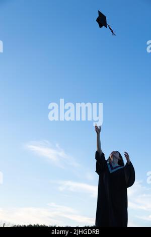 Junge Frau in Abschlusskleid, die ihre Mütze und ihr Mörtelbrett in die Luft wirft, mit einem blauen Himmel im Hintergrund Stockfoto