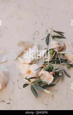 Sommerhochzeit florale Komposition. Nahaufnahme von Aprikosen-Oleanderblüten, Blumen und grünen Olivenbaumbästen. Beigefarbener Marmorhintergrund mit Seide Stockfoto