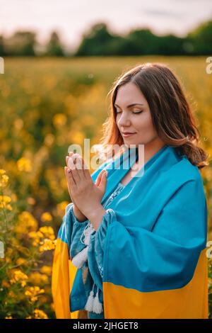 Ukrainische Patriotin, die mit Nationalflagge im gelben Rapsfeld betet. Betet, steht mit der Ukraine. Frieden, Unabhängigkeit, Freiheit, Sieg im Krieg. Stockfoto