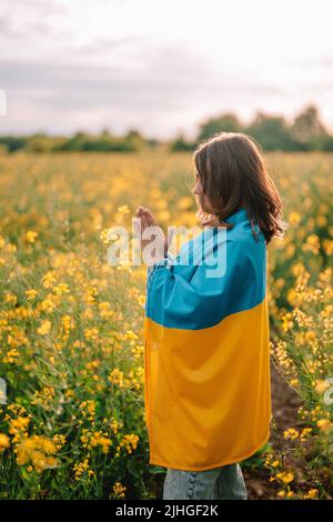 Ukrainische Patriotin, die mit Nationalflagge im gelben Rapsfeld betet. Betet, steht mit der Ukraine. Frieden, Unabhängigkeit, Freiheit, Sieg im Krieg. Stockfoto