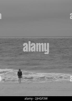 Eine einsame Frau am Strand hielt morgens ein Glas in der Hand, während sie auf das Meer starrte. Stockfoto