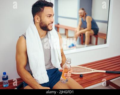 Junger Spieler trinkt Wasser, während er mit seinem Freund spricht. Zwei Freunde haben sich nach einem Squash-Match zusammengeschlossen. Zwei Männer, die nach einem Spiel in einem Umkleideraum im Fitnessstudio sitzen Stockfoto