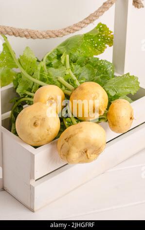 Frische gelbe Rübe mit Stielen und grünen Blättern in einer Schachtel auf weißem Holzgrund. Stockfoto