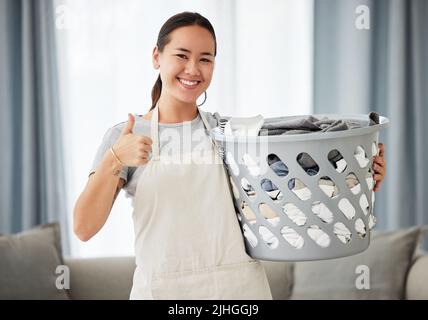 Porträt einer schönen jungen Frau mit gemischter Rasse, die beim Putzen ihrer Wohnung den Daumen nach oben zeigt. Eine glückliche asiatische Frau lächelt beim Falten der Wäsche Stockfoto