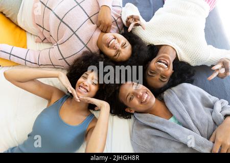 Hochwinkelporträt glücklicher birazialer junger Freundinnen, die gestikulieren und liegen, während sie sich auf dem Bett entspannen Stockfoto