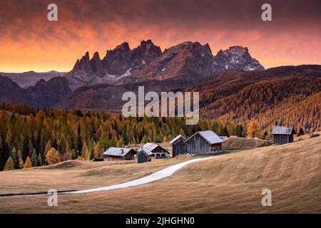 Unglaublicher roter Sonnenuntergang im Fuchiade-Tal in der Landschaft der italienischen Dolomiten. Holzhütten, orangefarbene Lärchen Wald und schneebedeckte Berge Gipfel im Hintergrund. Dolomiten, Italien Stockfoto