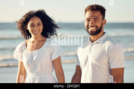 Glückliches gemischtes junges Paar, das sich die Hände hält, während es gemeinsam am Strand spazieren geht. Hispanische Paare reisen und genießen den Urlaub und sind romantisch auf Stockfoto
