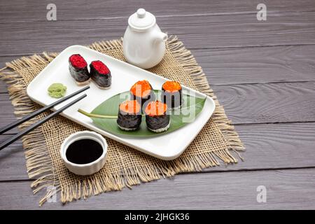 Nori Sushi mit Kaviar, schwarze Essstäbchen auf weißem Teller. Sojasauce in einer Schüssel. Boot mit weißer Soße. Quappe Serviette. Speicherplatz kopieren. Dunkler Holzhintergrund. Obere V Stockfoto