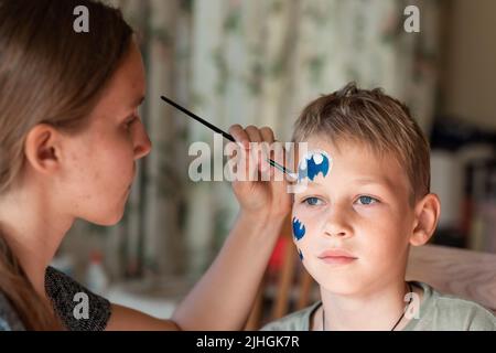 Kind Animator, Künstler Hand zeichnet Gesicht Malerei zu kleinen Jungen. Kind mit lustigen Gesichtsbemalung. Der Maler macht eine blaue batman-Maske auf das Gesicht des Jungen. Gesicht pa Stockfoto