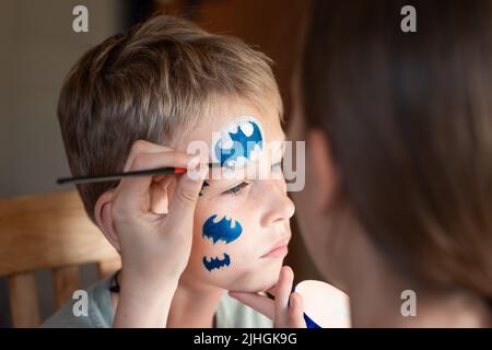 Kind Animator, Künstler Hand zeichnet Gesicht Malerei zu kleinen Jungen. Kind mit lustigen Gesichtsbemalung. Der Maler macht eine blaue batman-Maske auf das Gesicht des Jungen. Gesicht pa Stockfoto