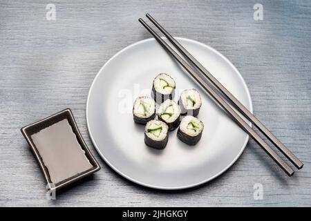 Sushi-Nori-Rollen und Essstäbchen auf blauem Teller. Saucenboot in schwarzer Schüssel. Flach liegend. Speicherplatz kopieren. Blauer Hintergrund Stockfoto