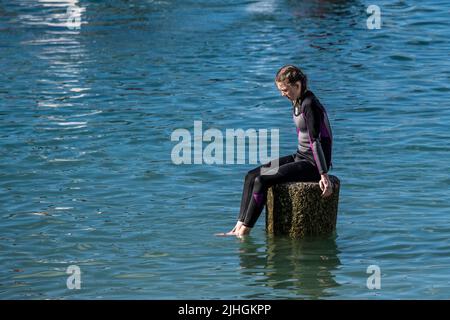 Eine junge Teenager, die bei Flut allein auf einem Granitbollard im Meer im Newquay Harbour in Cornwall im Vereinigten Königreich sitzt. Stockfoto