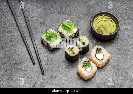 Sushi-Set mit Algen, schwarzem Sesam und Gurke. Essstäbchen, Wasabi in schwarzer Schüssel. Draufsicht. Schwarzer Hintergrund Stockfoto