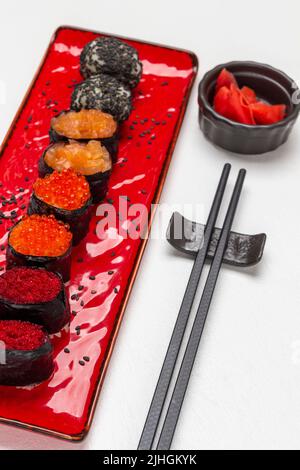 Set Sushi auf rotem Teller. Rosafarbener Ingwer in schwarzer Schüssel. Essenssticks. Draufsicht. Weißer Hintergrund Stockfoto