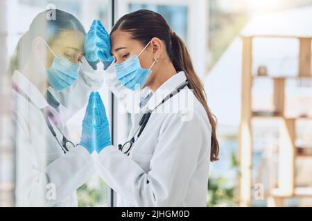 Ein junger gestresster und überarbeiteter Arzt trägt Maske und Handschuhe, während er an einem Fenster in einem Krankenhaus oder einer Klinik steht. Ein Weibchen sieht nur besorgt aus Stockfoto